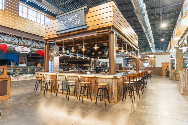 The Sawgrass Bar is photographed in the brightly lit Sawgrass Market. Numerous light fixtures hang from the edge of the bar structure and chairs neatly wrap around. At the heart of the market, the butcher shop and coffee shop can be seen in the background.